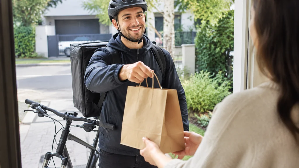 Delivery courier in a helmet handing a paper takeaway bag to a customer